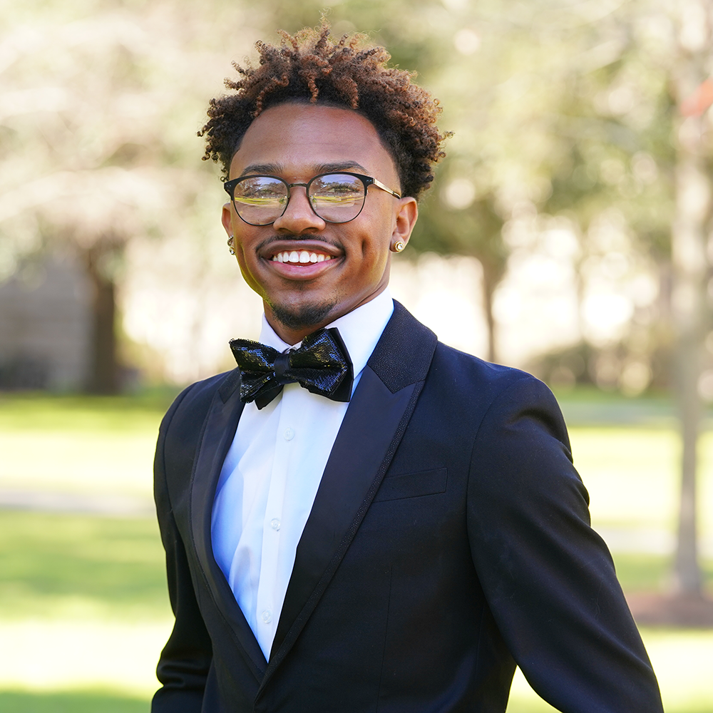 Smiling man in a black tuxedo, bow tie, and glasses outdoors.
