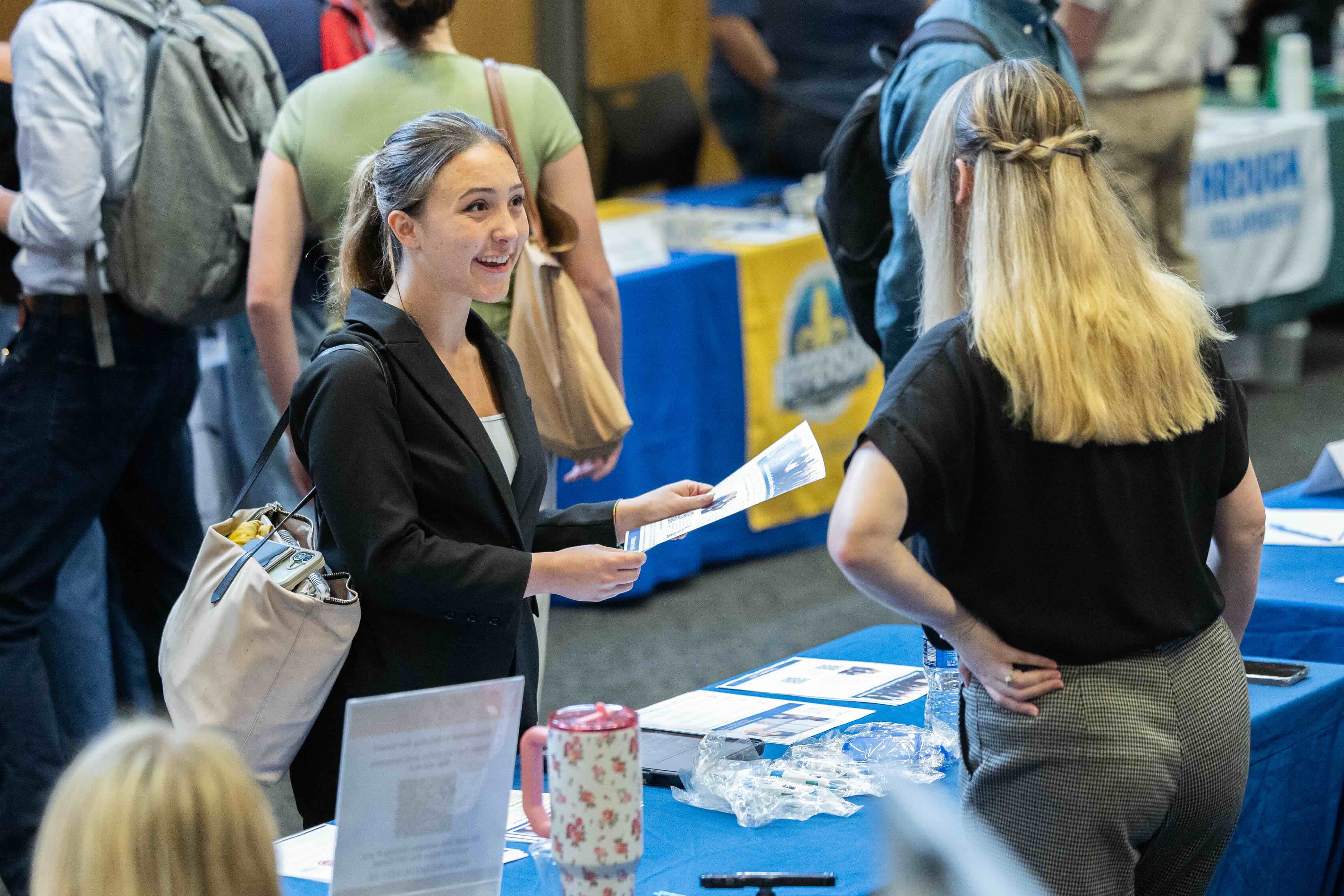 Smiling woman in suit jacket talks to another woman at a busy career fair.
