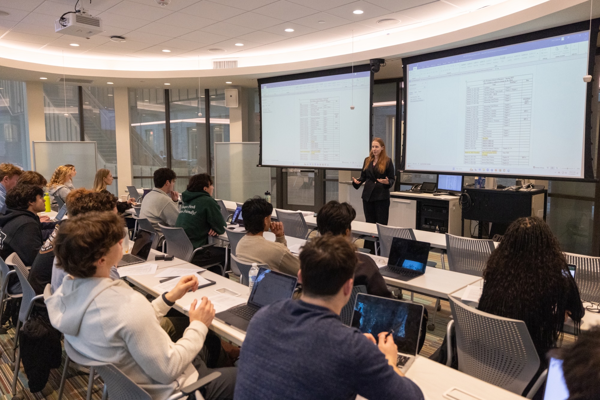 A woman presents data on two large screens to a class of students in a modern classroom.