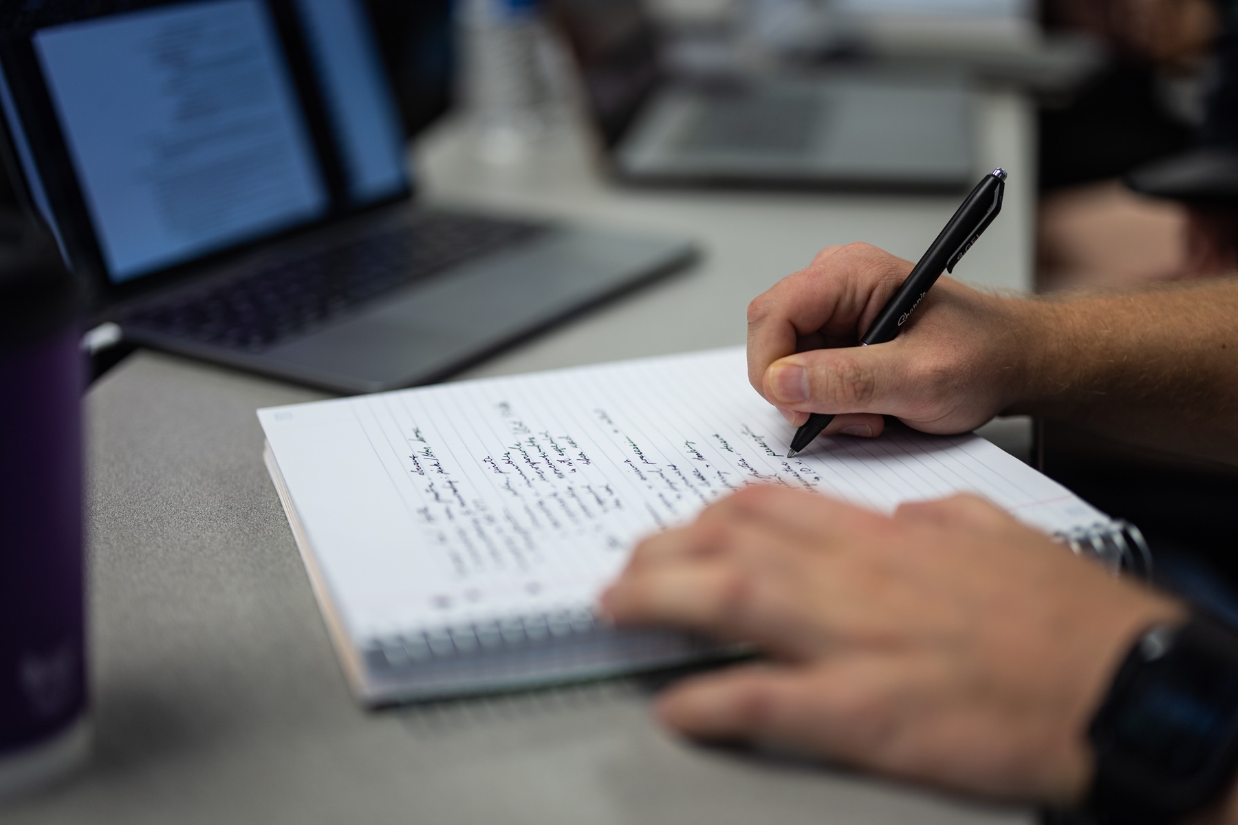 Person writing notes in a spiral notebook with a pen; laptop blurred in background.