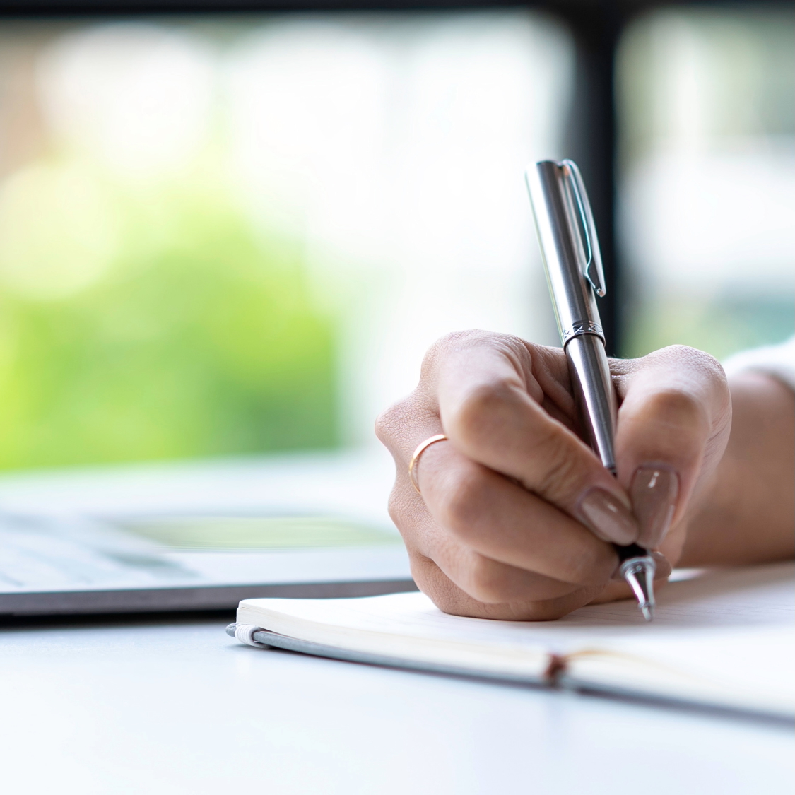Person's hand writing with a pen in a notebook, blurred laptop and greenery in background.