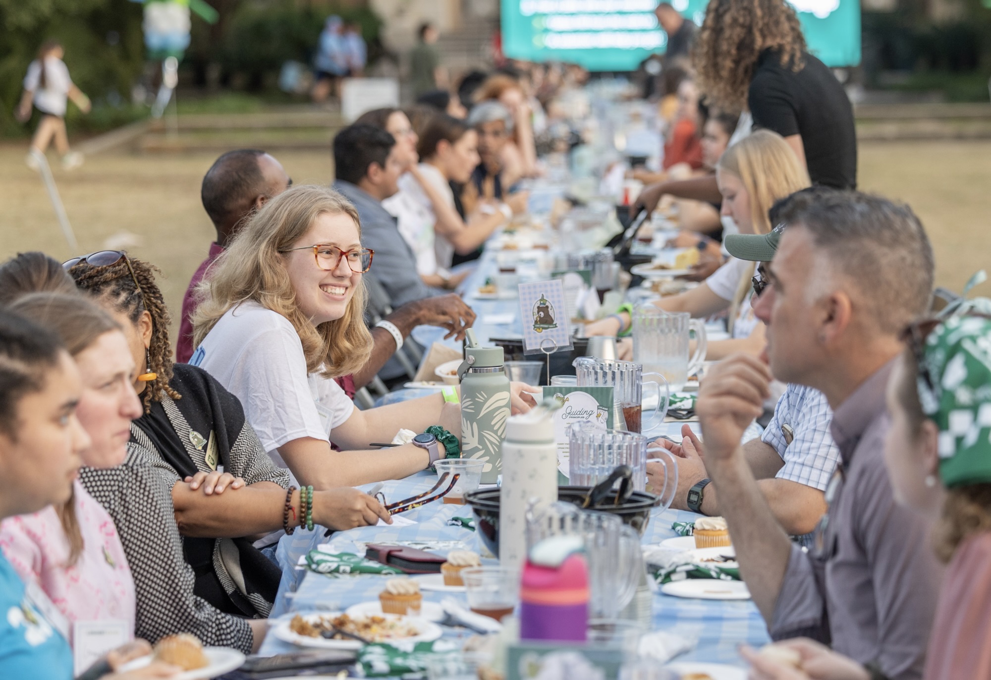 Smiling woman at a long outdoor table with many people dining outdoors.