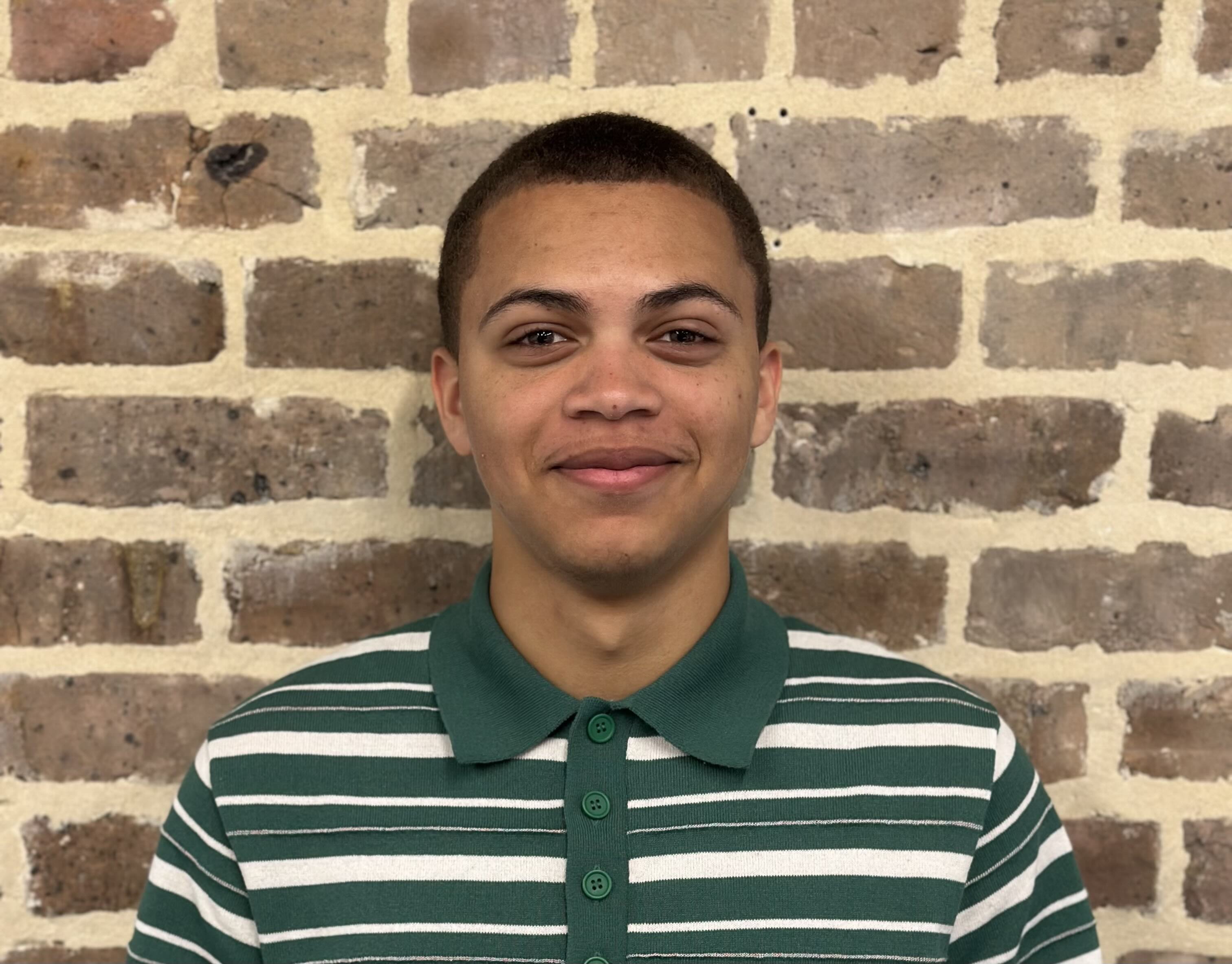 Young man with short hair, smiling in a striped polo shirt against a brick wall.