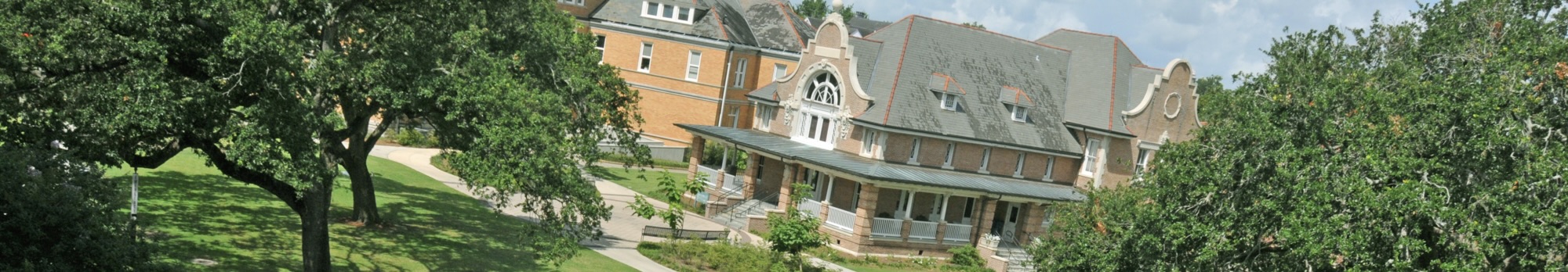 Large old building with an ornate roof, surrounded by lush green trees and a sloping lawn.