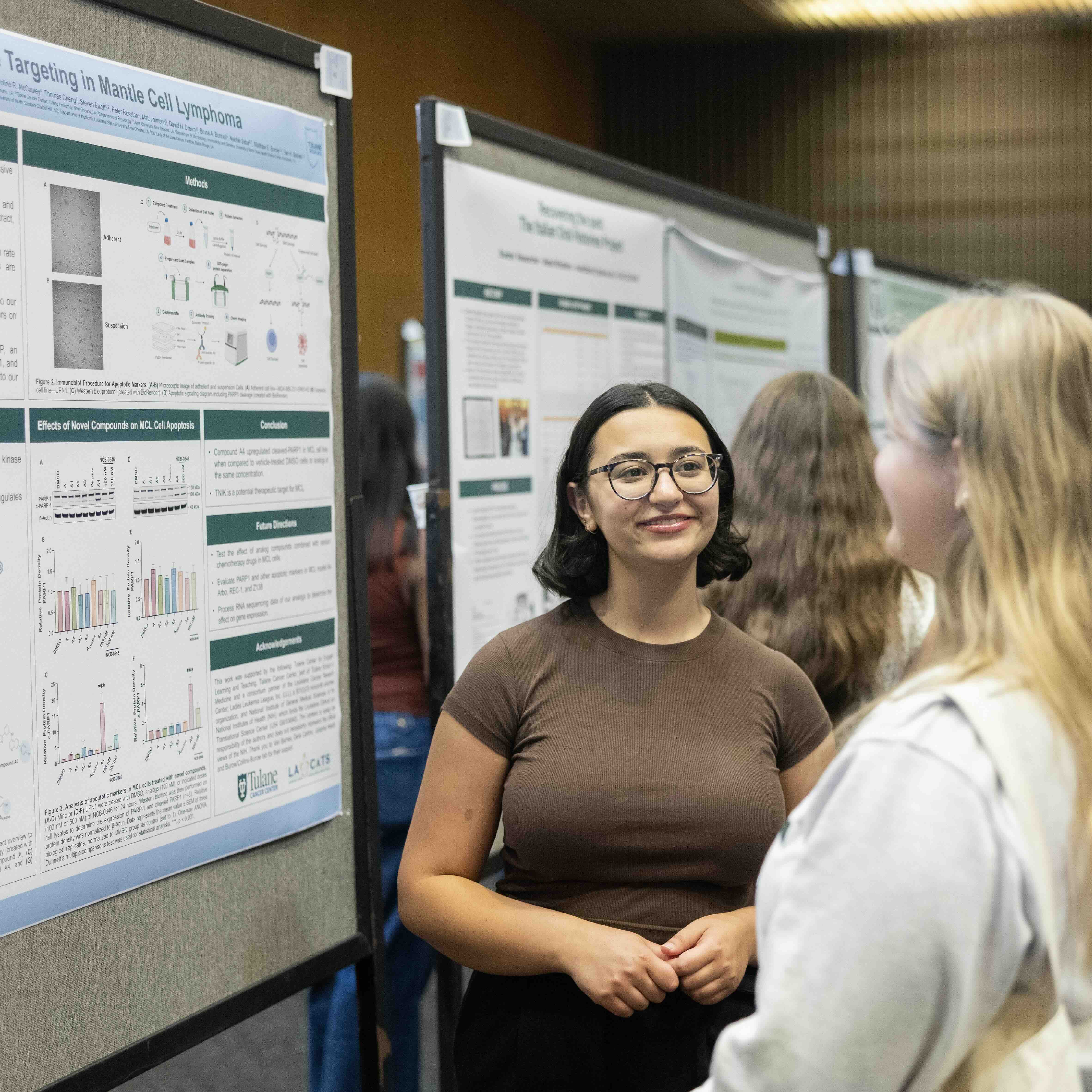 Smiling woman with glasses talks to another at a scientific poster session.