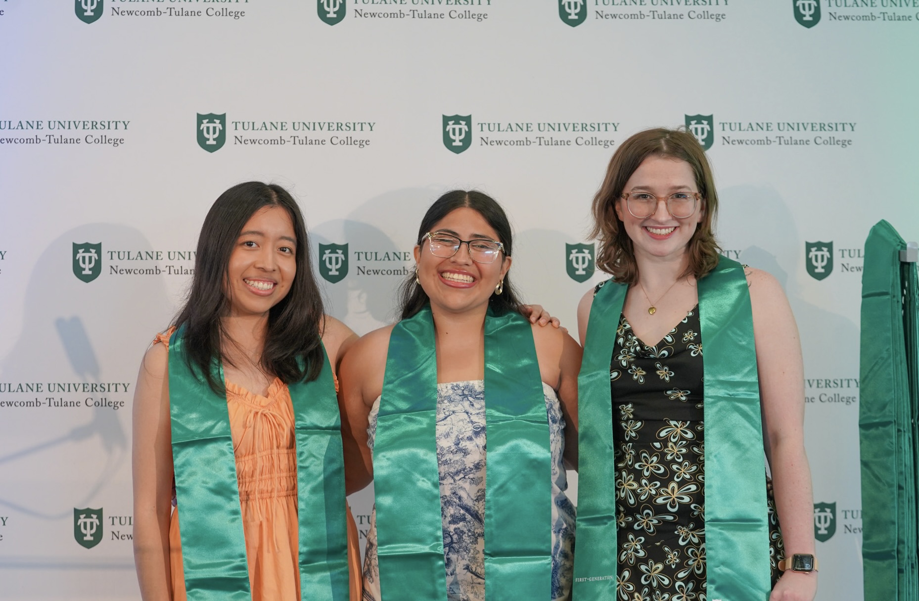 Three smiling women in green sashes stand in front of a Tulane University logo wall.