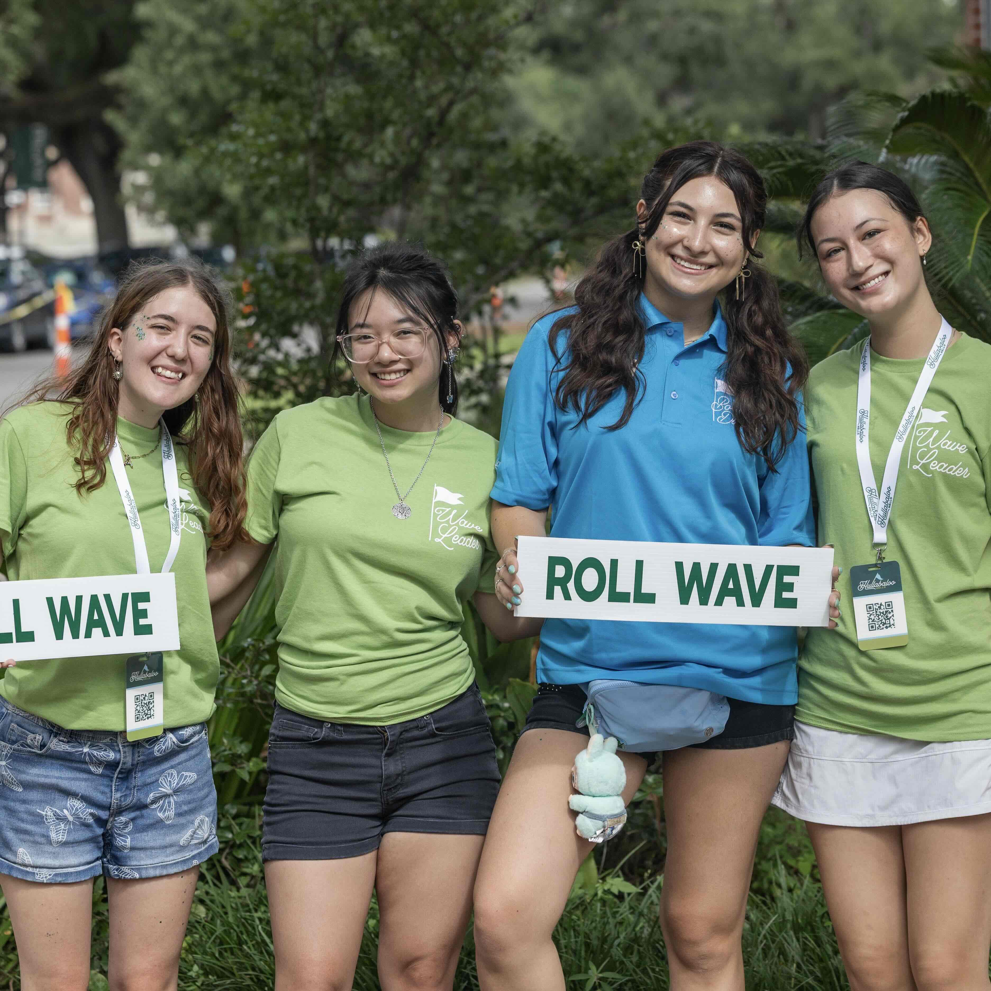 Four smiling young women hold "ROLL WAVE" signs outdoors.