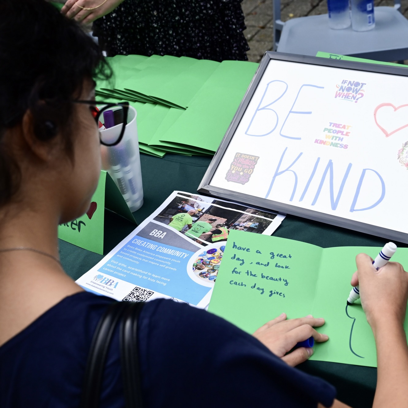Woman writes on green paper at a kindness booth, "BE KIND" sign in the background.