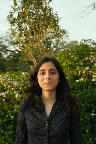 Young woman with dark hair, dark shirt, standing outdoors with green trees and foliage.