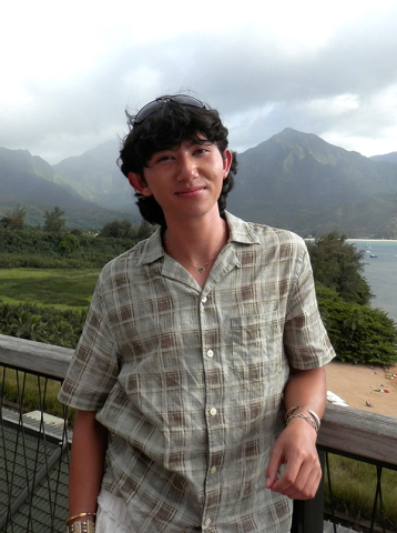 Smiling man in plaid shirt against a backdrop of green mountains and a coastal bay.